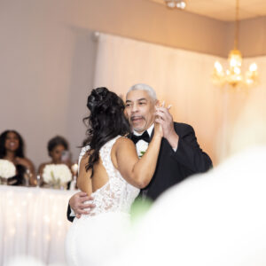 Bride dances with her father at a wedding reception. They are smiling and embracing on the dance floor beneath a chandelier with guests seated in the background.
