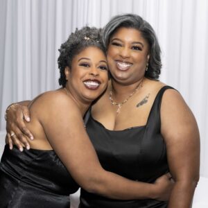 Two women wearing elegant black dresses smiling and embracing at a formal event standing in front of white draped curtains conveying a joyful and celebratory atmosphere.