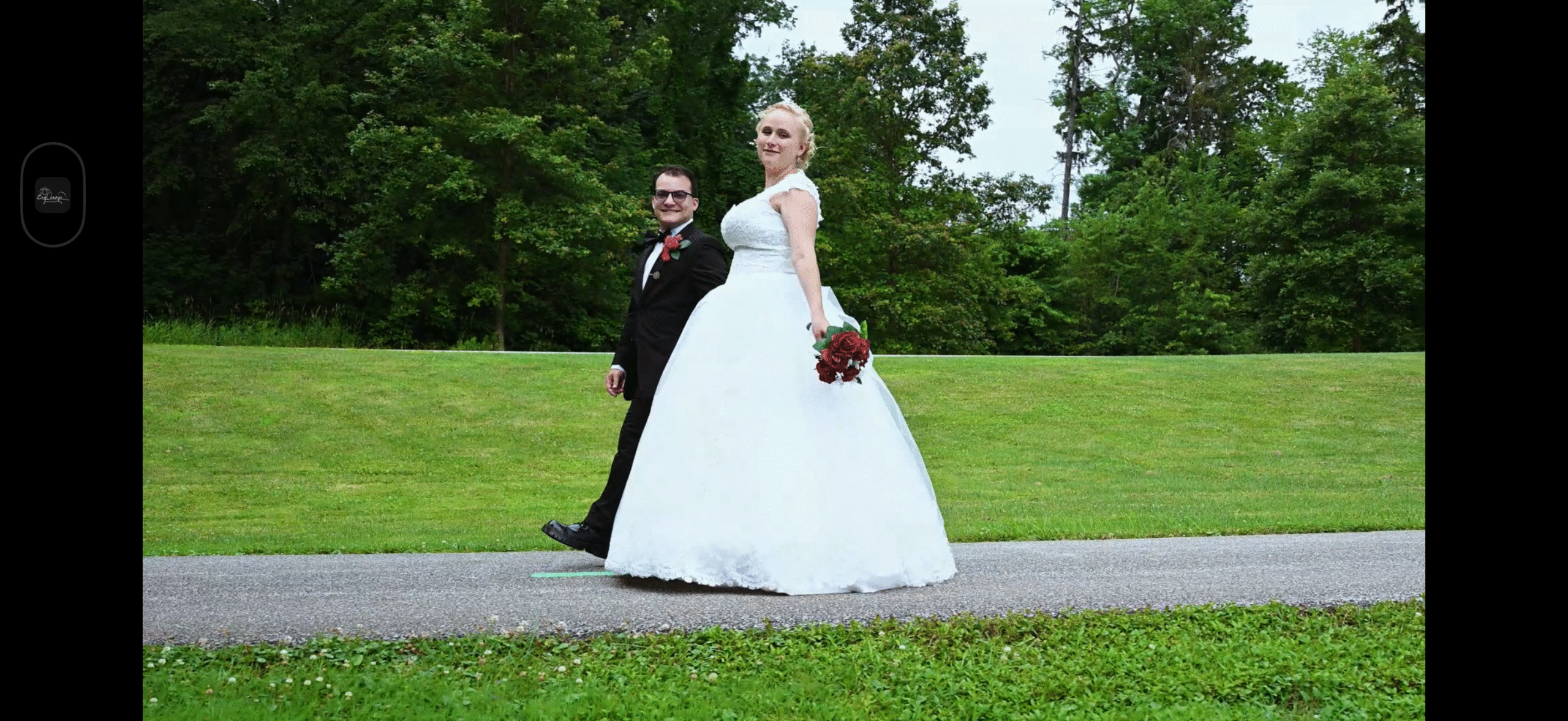 Bride and groom walking on a path in a lush green park with trees in the background the bride is wearing a white gown and holding a bouquet the groom is in a suit