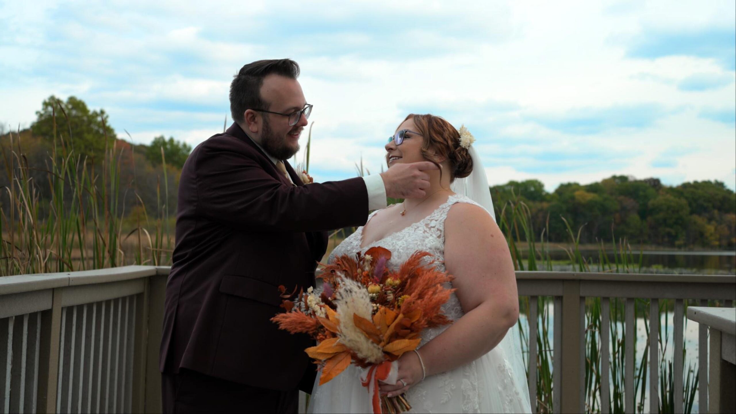 Bride and groom share a tender moment on a wooden deck with a lake and autumn trees in the background. The bride holds a vibrant bouquet of fall flowers.