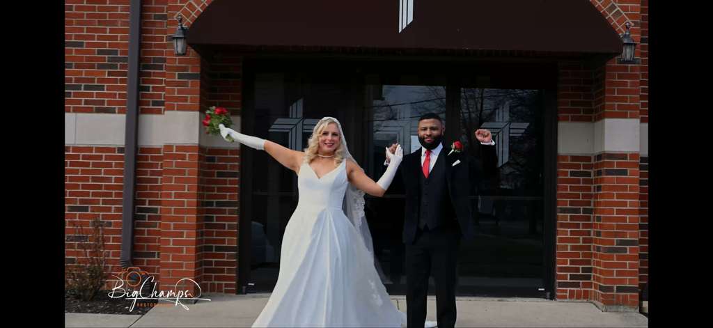 Bride and groom celebrating outside a brick building on their wedding day. The bride holds a bouquet and smiles, while the groom raises his arm in joy.