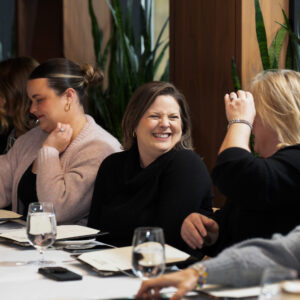 A group of women enjoying a lively conversation at a dining table set with menus and wine glasses in an elegant restaurant setting with natural lighting and decor.