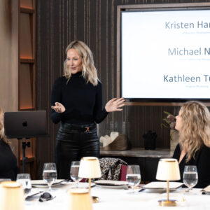 A woman gives a presentation to an attentive group in a modern conference room with a digital display in the background. Elegant table setting with glasses and lamps.