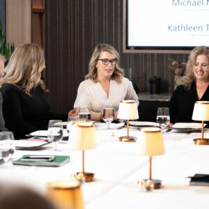 A group of people engaged in a professional meeting around a table with documents and glasses. A presentation screen is visible in the background displaying names.