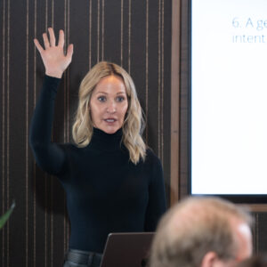 Corporate presenter speaking confidently during a professional business meeting, wearing a black outfit and gesturing while addressing the team