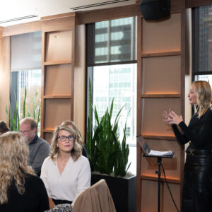 A woman stands and presents to an attentive audience in a modern conference room filled with natural light and tall green plants along the windows.