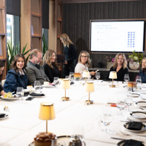 A diverse group of people sits around a long conference table engaged in a meeting. A presentation screen is visible, along with elegant table settings and soft lighting.