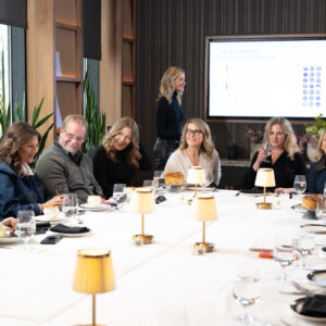 A group of people engaged in a business conference at a large table with a presentation screen in a modern room adorned with plants and lamps.