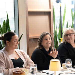 Three women seated at a restaurant table enjoying a conversation. The scene features elegant lighting and lush green plants, creating a warm and inviting atmosphere.