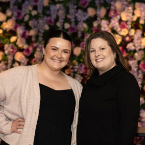 Two attendees posing in front of a colorful floral backdrop at a corporate event.