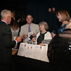 A group of people engaged in conversation at a formal event standing by a table with documents. They are dressed in evening attire, holding drinks and smiling warmly.