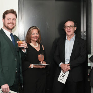 A group of people in formal attire enjoying drinks at a social event. They are smiling and standing together in a room with an elevator in the background.