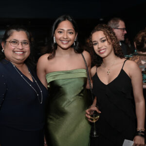 A group of three women smiling at a formal event, wearing elegant attire with others socializing in the background, creating a friendly and celebratory atmosphere.