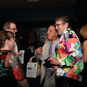People socializing at a vibrant event, engaging in conversation and laughter. A man in a colorful shirt stands out among the group, creating a lively atmosphere.