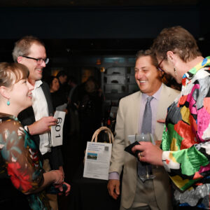 A group of four people enjoying a lively conversation at a social event. They are dressed in colorful and formal attire and appear to be holding drinks and smiling.