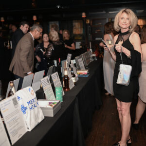 Guests exploring a display of items at a formal event, with a woman in a black dress holding a brochure. The room is elegantly decorated for an evening gathering.