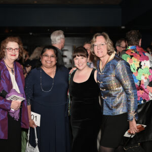 A group of four women smiling and posing at a social event with a busy background filled with other attendees creating a lively and festive atmosphere.