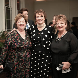 Three women dressed elegantly in a formal event setting smiling at the camera with other guests in the background enhancing the atmosphere of a sophisticated gathering.