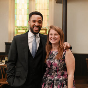A man in a suit and a woman in a floral dress smile at the camera. They are standing in a warmly lit room with wooden chairs and set tables in the background.