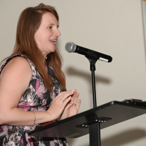 Woman speaking at a podium during a professional event in a floral dress with a microphone on the stand and a partially visible banner in the background.
