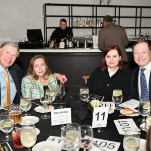 A group of four people at a formal dinner event sitting at a round table with drinks and menus in a sophisticated restaurant setting with a bartender in the background.