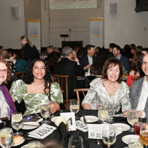 Group of people seated at a round table during a formal event surrounded by others in a large hall with dim lighting and elegant decorations.