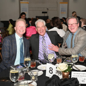 Three men in suits smiling at a formal gathering seated around a table with drinks and brochures with a crowd in the background and soft lighting creating a warm ambiance
