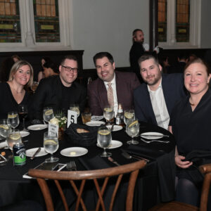 A group of people sitting around a table at an elegant indoor event. The setting features dim lighting, wooden chairs, and a table set with plates and glasses.