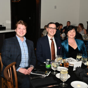 Three people smiling and sitting at a dinner table during a formal event in a restaurant setting with plates and drinks on the table.