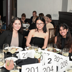 A group of women enjoy a formal dinner event seated at a table with white place cards and plates of food surrounded by attendees in a warmly lit dining area.