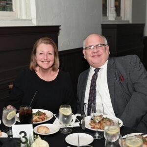 A smiling couple enjoying a formal dinner at a restaurant with elegantly set tables featuring plates of food and glasses of water and wine creating a warm and inviting atmosphere
