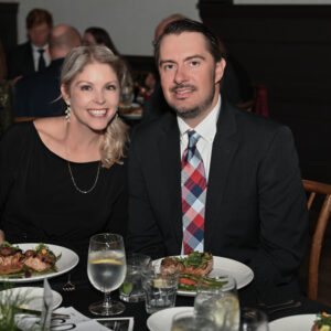 Smiling couple enjoying a formal dinner at an elegant event with plates of gourmet food and glasses of water on a beautifully set table in a sophisticated setting.