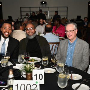 Group of four men smiling at a formal dinner event sitting around a table with drinks and plates in a warmly lit room with other guests and table number cards visible.