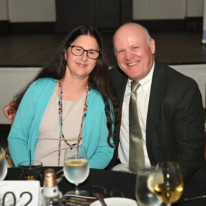 Smiling man in suit and woman with long hair and glasses seated at a table with water and wine glass at formal event in elegant venue.
