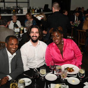 Three people enjoy a formal dinner at a lively restaurant event. They are smiling at the camera, surrounded by elegantly set tables and other guests dining in the background.