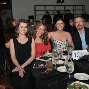 A group of people sitting at a restaurant table enjoying a social gathering with plates of food and drinks surrounded by other guests in a dimly lit event space.