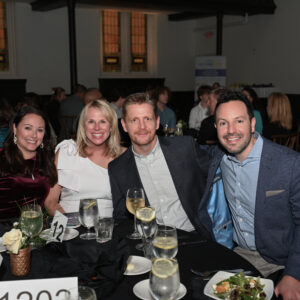 Group of four people smiling at a formal dinner event, seated around a table with numbered place cards and elegantly set dinnerware in a dimly lit room.