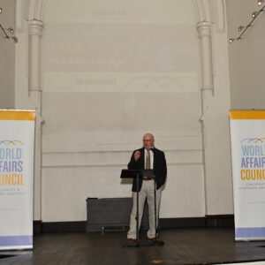 Speaker at World Affairs Council event standing on stage with banners on either side delivering a speech at a podium in a well-lit room with high ceilings.