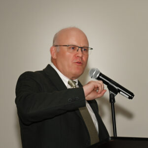 A speaker in a suit and glasses delivers a presentation at a podium with a microphone against a neutral background, highlighting public speaking and professional communication.