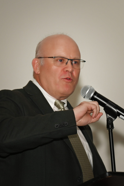 A speaker in a suit and glasses delivers a presentation at a podium with a microphone against a neutral background, highlighting public speaking and professional communication.