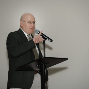 Man in black suit jacket and tie speaks into a microphone at a podium during a conference. A banner with text is partially visible on the right side of the image.