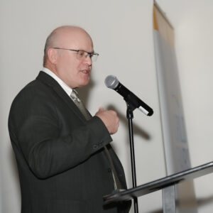 Man speaking at a podium with a microphone at a formal event wearing a suit and tie delivering a speech in a well-lit conference room with a white backdrop.