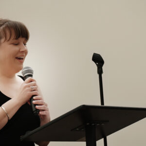A woman in a black dress is speaking into a microphone at a podium in a well-lit room. She seems confident and engaged, suggesting a presentation or speech setting.