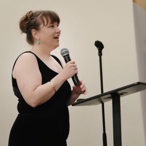 A woman in a black dress speaks confidently into a microphone at a podium set against a plain background. She appears to be presenting at an event or conference.