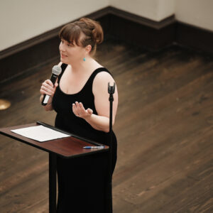A woman in a black dress speaks confidently into a microphone standing at a podium on a wooden stage delivering a presentation or speech to an audience.