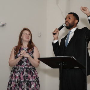 Two people standing on stage during a presentation. A woman in a floral dress is looking at a man in a suit who is speaking into a microphone at a podium.