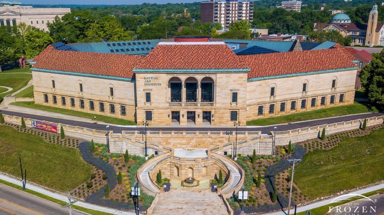 Aerial view of an elegant stone building with a red roof surrounded by greenery and walkways in a vibrant urban setting on a sunny day.