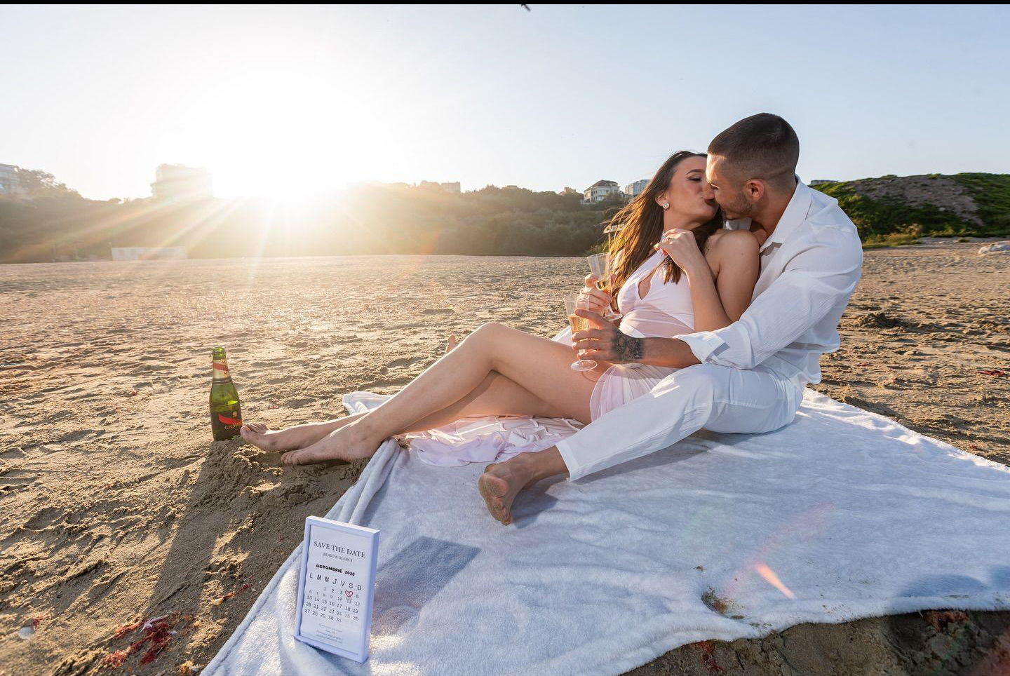 Couple in white outfits sharing a romantic moment on a beach blanket at sunset Champagne bottle nearby Sunny seashore background perfect for romantic getaways or celebrations