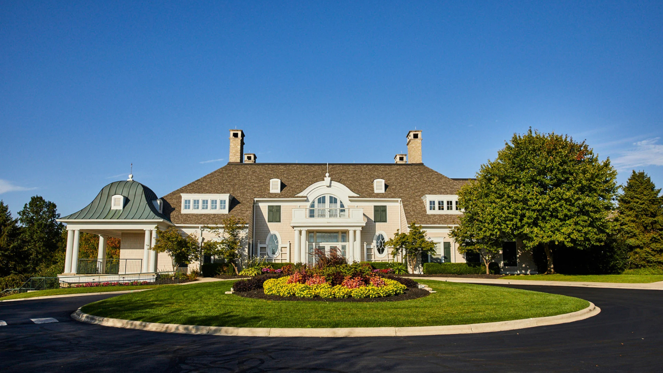 Elegant country estate with a well-maintained circular driveway surrounded by lush greenery and colorful flower beds against a clear blue sky. Luxury residential architecture.