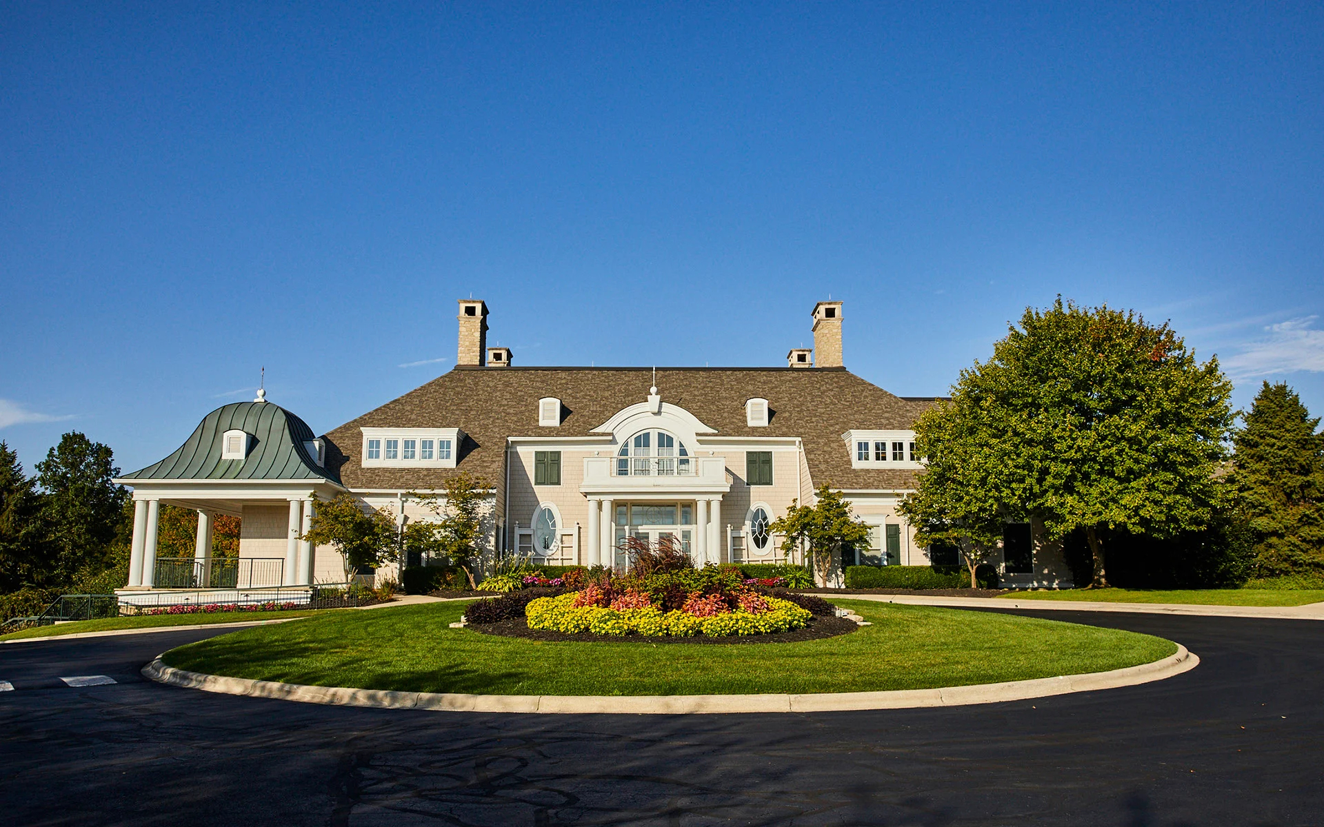 Elegant country estate with a well-maintained circular driveway surrounded by lush greenery and colorful flower beds against a clear blue sky. Luxury residential architecture.